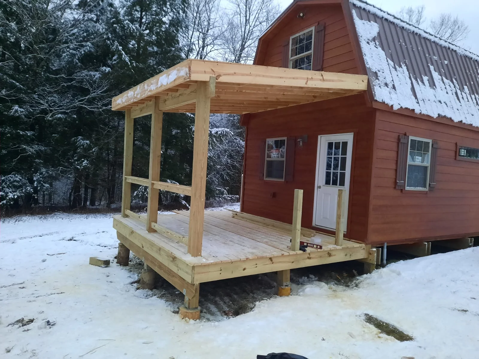 Wooden covered porch with gable roof attached to red cabin, pressure-treated lumber framing and decking
